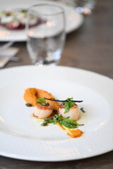 Close-up view of a gourmet appetizer with seared scallops served on a white plate in a fine dining restaurant. Elegant presentation with herbs, sauce, and pur&eacute;e highlights modern European cuisine and 