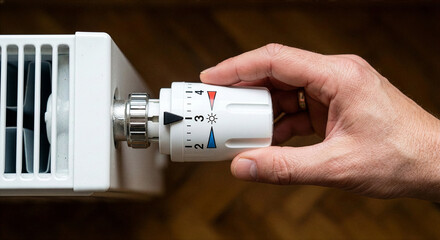 A hand turning the white thermostat dial of a modern household radiator to adjust the temperature.