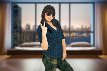 A woman is posing in a room with a city view in the background. She is wearing a blue shirt and green pants