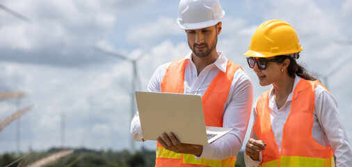 Engineer wearing safety vests and helmets analyzing wind energy data on laptop at wind farm, representing teamwork and sustainable technology development.