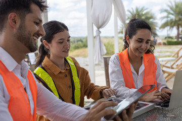 Team of engineers collaborating with digital devices at outdoor office table, planning renewable energy project with wind turbines in background, showcasing teamwork and green technology.