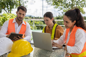 Team of engineers collaborating with digital devices at outdoor office table, planning renewable energy project with wind turbines in background, showcasing teamwork and green technology.