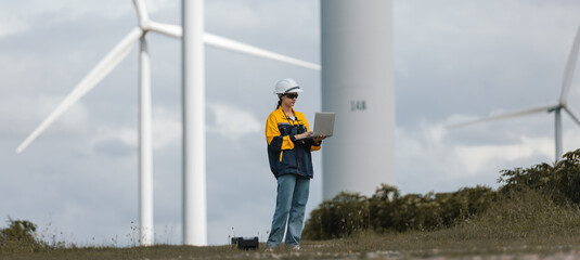 Female wind turbine technician standing with laptop and toolbox at wind farm, representing renewable energy maintenance, green technology, professional engineering in clean power.