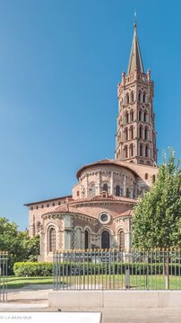 The cobbled parvis of the Basilica of Saint Sernin in Toulouse, France, timelapse hyperlapse. Largest romanesque church in Europe with brick bell tower, arched doors, green trees and blue sky