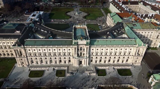 Aerial drone shot panning over Hofburg Palace from behind, facing the palace grounds in Vienna, showing the surrounding cityscape, winter sunlight, cloudy skies, and tourists walking in the grounds.