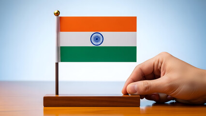A hand holds a miniature Indian flag on a wooden base against a bright background