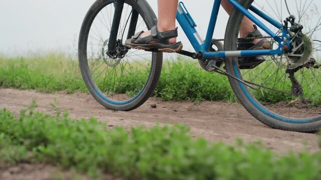 Cyclist pedaling on dirt trail, blue bike closeup showing feet in sandals, rear wheel kicking up soil, visible chain and gears, grassy field background, overcast sky, sense of motion and outdoor
