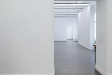 A view looking into an empty, spacious room with white walls and a gray tiled floor. The ceiling features rows of industrial fluorescent lights, and the space connects to another room.