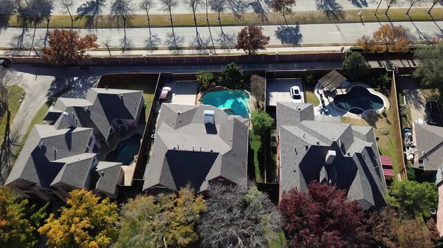 Divided four-lane road flanked by fenced suburban homes and landscaped sidewalks in Coppell, Texas. Autumn trees line the median, upscale kidney-shaped pool and shaded patio visible in backyard