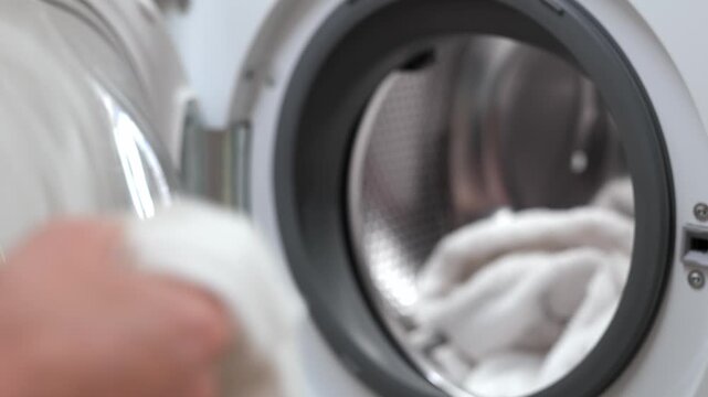 Caucasian woman places a white towel into a washing machine, then adds more laundry, demonstrating the process of loading laundry for washing in a modern laundry room
