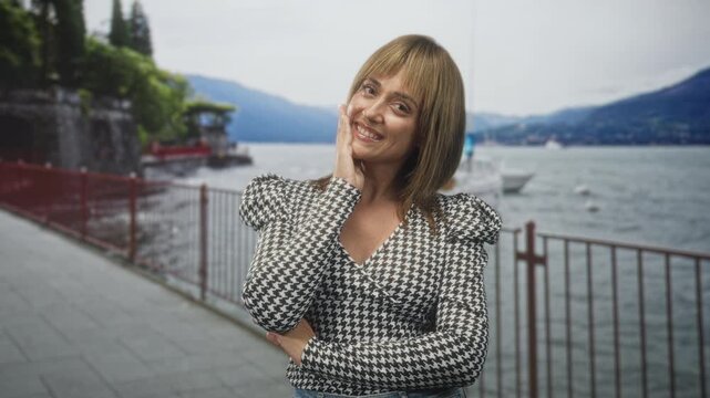 Woman touching cheek while smiling on a lakeside promenade beside a metal railing and moored boats, wearing houndstooth patterned top and jeans; joy.