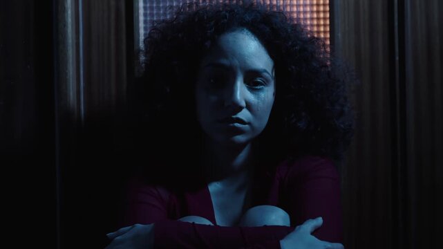 In a dark home environment, a heartbroken young Latina woman sits on the floor and looks directly into the camera, expressing pain, vulnerability, and emotional exhaustion.