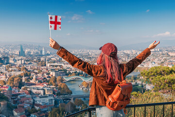 Cheerful woman holding a Georgia flag enjoying the panoramic view of Tbilisi cityscape and the Kura River © EdNurg