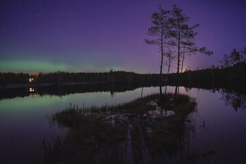 Peaceful Northern Lights over a calm lake with silhouettes of trees and a remote cabin in Finland.