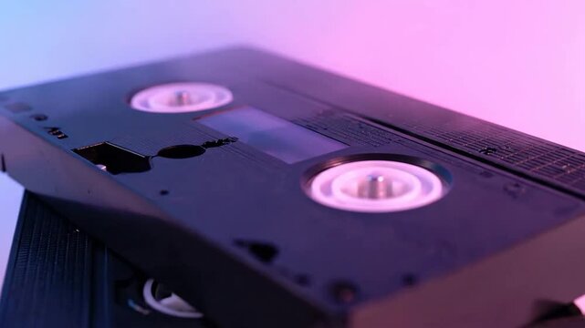 Stacked Black VHS Tapes Under Ambient Lighting with Pink and Blue Hues on White Backdrop