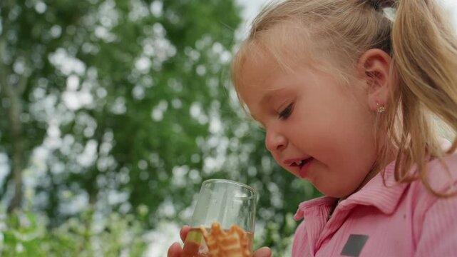 caucasian girl eating wafer cookie outdoors, holding glass jar of honey, curious tasting and gentle nibbling, sticky fingers, pink shirt with ponytail, soft sunlight filtering through green trees,