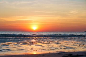 Golden sunset over a calm ocean with the sun touching the horizon, warm orange sky reflected on wet...