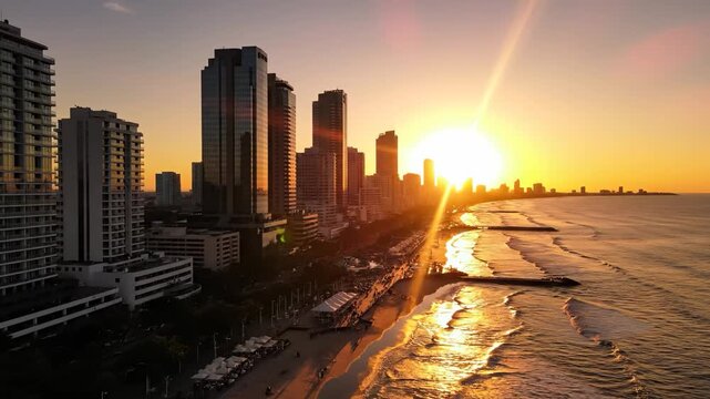 A beautiful sunset illuminates the city skyline, casting a warm glow over the ocean waves. The reflective buildings and sandy beach enhance the vibrant sunset and coastal scene.
