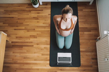 Pregnant woman practicing yoga online at home with laptop.