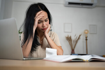 An Asian woman sits indoors holding receipts and a credit card, appearing concerned. The scene reflects financial stress, personal budgeting, and the emotional impact of managing expenses in a home