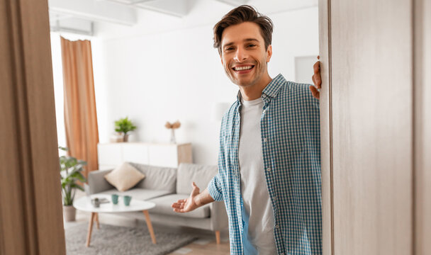 Portrait of cheerful man welcoming inviting visitor to enter his home, happy young guy standing in doorway of modern apartment, millennial holding door looking out showing living room with hand