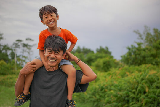 Smiling man giving a piggyback ride to a happy little boy outdoors with green foliage background. Joyful family bonding and playful childhood concept