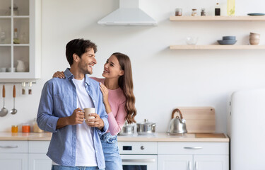 Cheerful millennial european wife hugs husband with cup of hot drink in minimalist kitchen...
