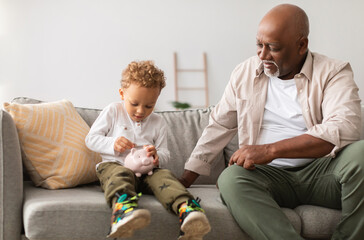 Happy African American Grandfather And His Grandson Putting Coin Money In Piggybank Sitting On Sofa...