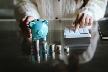 Blue piggy bank with stacked coins on a table, while a person uses a calculator in the background, representing saving money, budgeting, personal finance, and financial planning concepts.