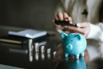 Blue piggy bank with stacked coins on a table, while a person uses a calculator in the background, representing saving money, budgeting, personal finance, and financial planning concepts.