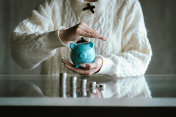 Blue piggy bank with stacked coins on a table, while a person uses a calculator in the background, representing saving money, budgeting, personal finance, and financial planning concepts.