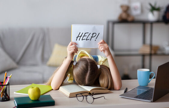 Learning Difficulties, Education Concept. Sad tired frustrated schoolgirl sitting at the table with book and laptop, holding paper with word Help. Overworked child asking for rest. Homeschooling