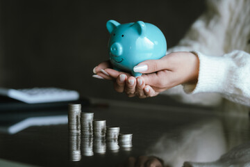 Blue piggy bank with stacked coins on a table, while a person uses a calculator in the background, representing saving money, budgeting, personal finance, and financial planning concepts.