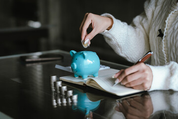 Blue piggy bank with stacked coins on a table, while a person uses a calculator in the background, representing saving money, budgeting, personal finance, and financial planning concepts.