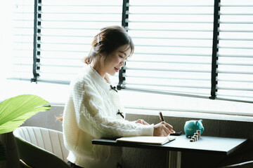 Young Asian woman calculating personal finances at home, using a calculator beside a piggy bank and coins on a desk by the window, symbolizing budgeting, saving money, and financial planning.