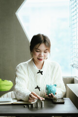 Young Asian woman calculating personal finances at home, using a calculator beside a piggy bank and coins on a desk by the window, symbolizing budgeting, saving money, and financial planning.
