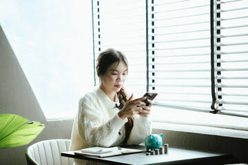 Young Asian woman calculating personal finances at home, using a calculator beside a piggy bank and coins on a desk by the window, symbolizing budgeting, saving money, and financial planning.