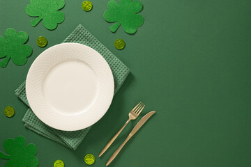 Festive St Patricks day table setting with white plate, gold cutlery and green shamrock decorations on green background. Elegant holiday mockup with copy space. View from above.