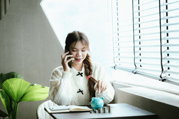Young Asian woman calculating personal finances at home, using a calculator beside a piggy bank and coins on a desk by the window, symbolizing budgeting, saving money, and financial planning.