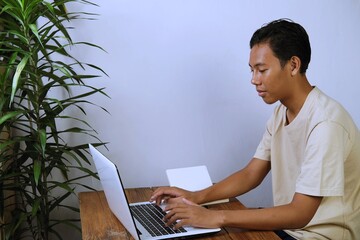 Young Asian man focused on working with a laptop at a simple home workspace. Modern daily life concept with copy space, technology, productivity, and remote work lifestyle.