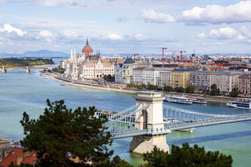 Obraz premium High angle panorama of trees, roofs and the 1849&nbsp;Széchenyi Chain Bridge over the Danube River and historic buildings including the Gothic Renaissance Parliament on the Pest side of the city, Budapest