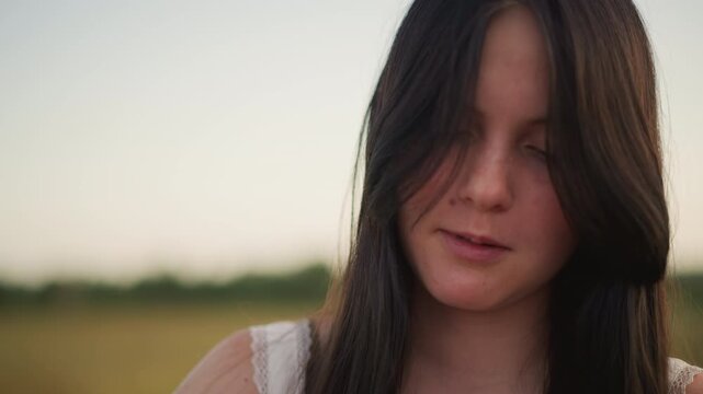 young woman in sunset field, soft golden light, long dark hair falling over face, gentle smile with closed eyes, candid headshot in meadow, summer dusk atmosphere, cinematic shallow depth, peaceful