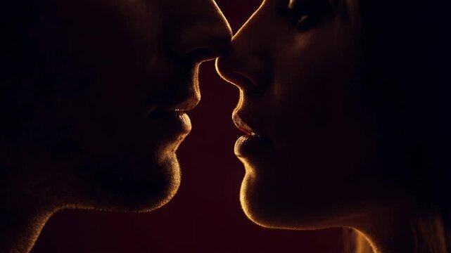Passionate couple in silhouette sharing an intimate moment, their profiles illuminated by a warm backlight as they gently get closer for a tender kiss against a dark red background