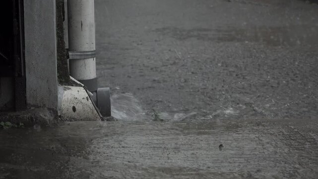 Heavy rain water pouring from a roof gutter pipe in front of a house, capturing intense rainfall and water drainage during a stormy weather condition.