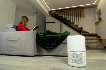 Woman relaxing on sofa with book in modern living room while air purifier improves indoor air quality, cozy evening home lifestyle, wellness, comfort and clean air indoors