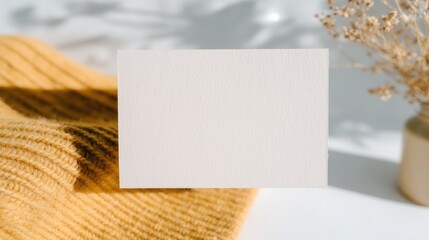 Blank white card mockup on cozy yellow knit sweater, with dried flowers in vase and soft light shadows. Ideal for wallpaper, poster, stationery design: minimalist warm aesthetic template for notes