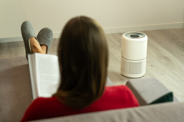 Woman relaxing on sofa with air purifier working in the living room, concept of clean indoor air, home comfort, air quality improvement and modern lifestyle. Stop smog and suspended dust 