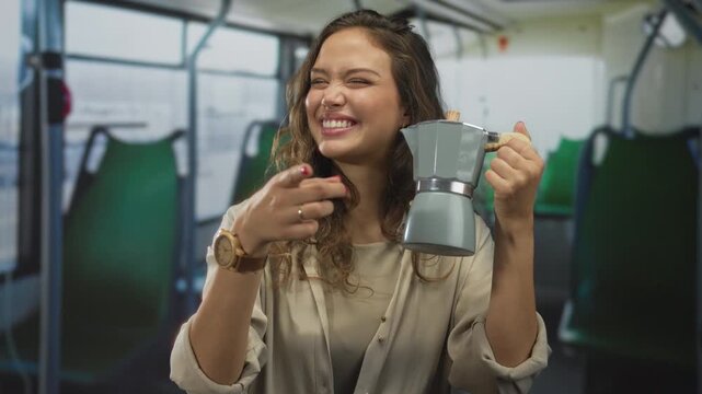 Young hispanic woman holds stovetop coffee maker and points finger to camera inside building; playfulness.