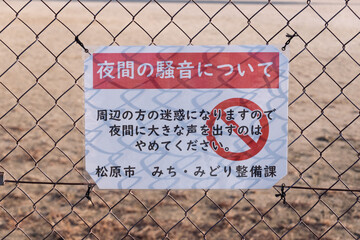 松原市民運動広場の夜間騒音警告：逆光で金網の影が文字に重なる「通行止め」マーク入りの看板