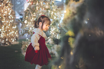 Adorable little Asian girl in a red velvet dress looking at a frosted Christmas tree decorated with warm bokeh lights during holiday celebration at home